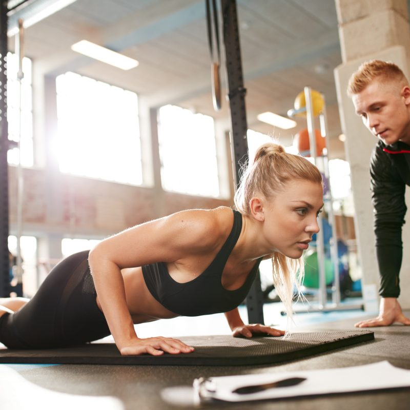 Indoor shot of young female exercising with personal trainer at gym. Fitness woman doing push ups with her personal trainer at health club.